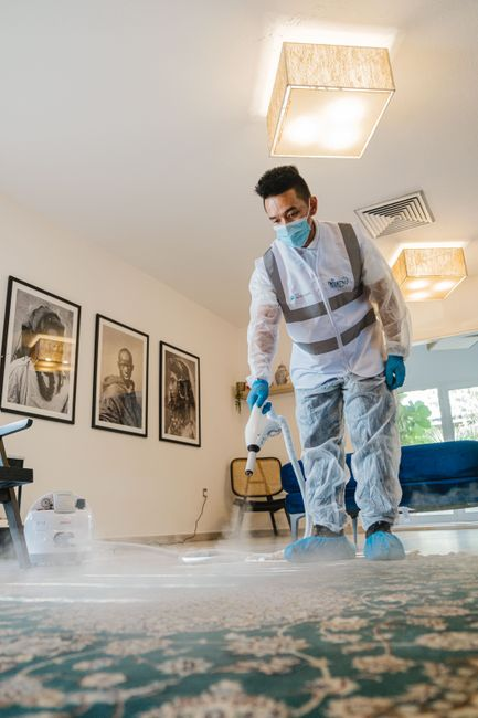 A person wearing gloves cleaning along a baseboard near a wall vent in a room.