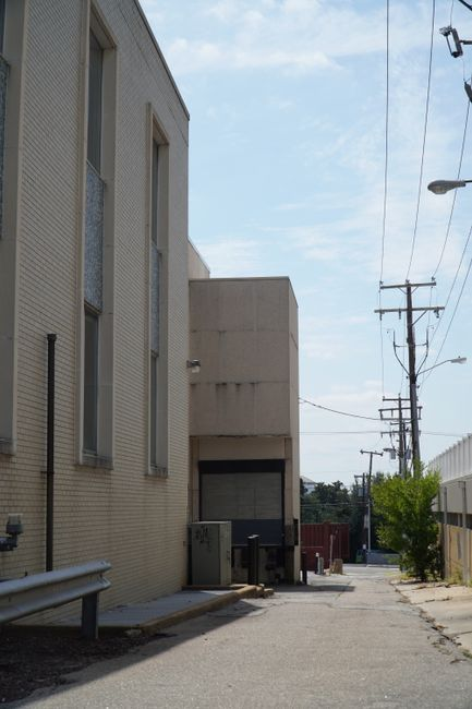 An exterior service edge with utility equipment, pipes, and a drainage route along a facility perimeter.