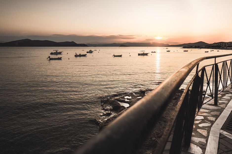 A lively Greek seaside promenade beside a broad beach at sunset, with cafes and people strolling near the water.